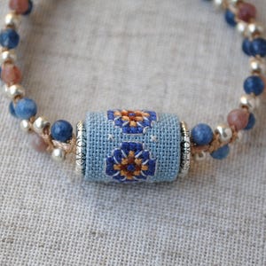 May include: A handmade bracelet featuring a central light blue cylinder with embroidered floral designs in blue and orange. The bracelet is adorned with blue, brown, and silver beads, strung on a beige cord. The design is close up and the background is a light beige fabric.