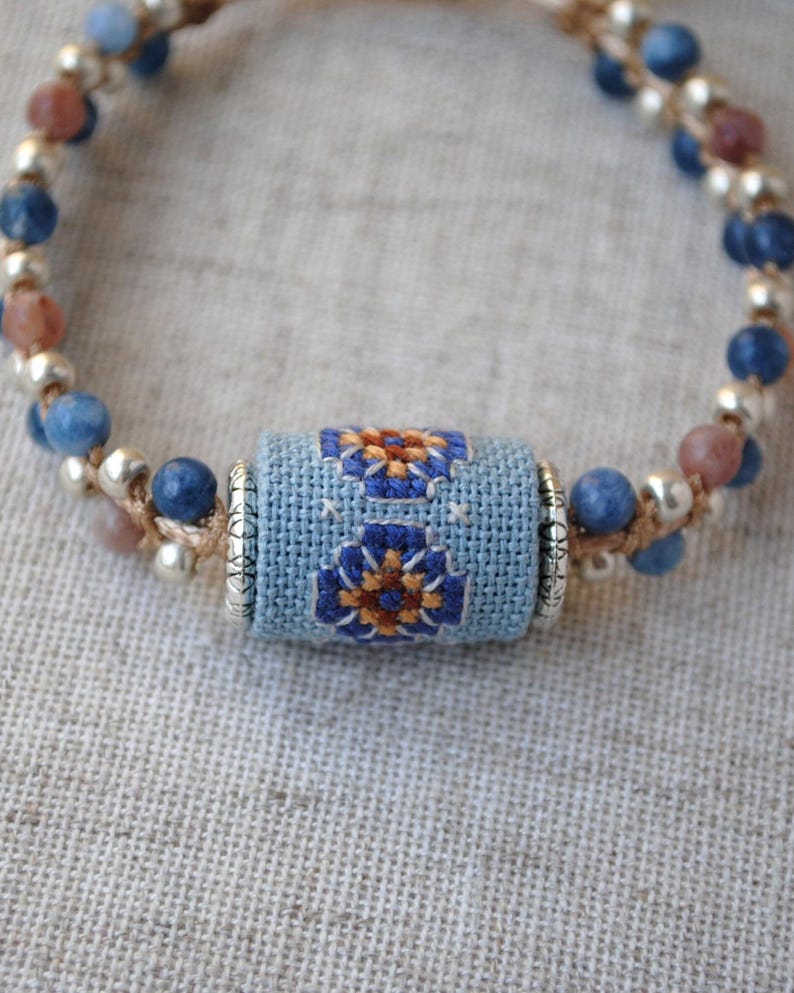 May include: A handmade bracelet featuring a central light blue cylinder with embroidered floral designs in blue and orange. The bracelet is adorned with blue, brown, and silver beads, strung on a beige cord. The design is close up and the background is a light beige fabric.