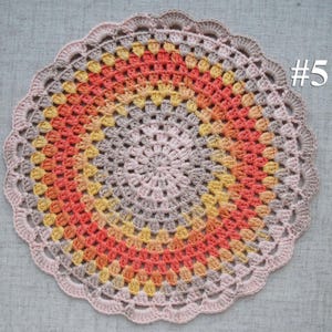 Puede incluir: Un tapete de ganchillo circular hecho a mano en tonos melocot&oacute;n, naranja, amarillo y gris, con un borde festoneado. El n&uacute;mero "#5" es visible en la esquina superior derecha. El tapete est&aacute; colocado sobre una superficie de tela gris.