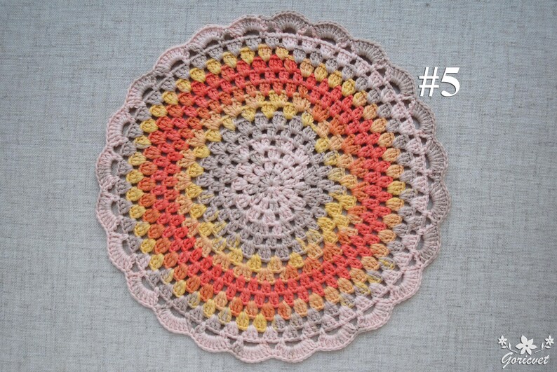 Puede incluir: Un tapete de ganchillo circular hecho a mano en tonos melocot&oacute;n, naranja, amarillo y gris, con un borde festoneado. El n&uacute;mero "#5" es visible en la esquina superior derecha. El tapete est&aacute; colocado sobre una superficie de tela gris.