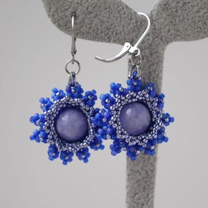 Op de afbeelding: Handgemaakte oorbellen met een centrale lavendelkleurige steen en een blauw kralenontwerp. De oorbellen hebben een bloemachtig patroon en zilverkleurige oorhaaksluitingen. De oorbellen worden tentoongesteld op een grijze oorbelstandaard.