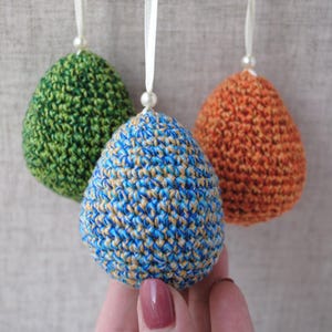Pu&ograve; includere: Tre uova di Pasqua all'uncinetto appese a un nastro bianco. Le uova sono verdi, blu e arancioni.