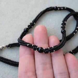 May include: A black beaded necklace with silver accents and a central black bead. The necklace is made of small, tightly woven beads, creating a textured appearance. The clasp is silver. The jewelry is displayed against a light gray background.