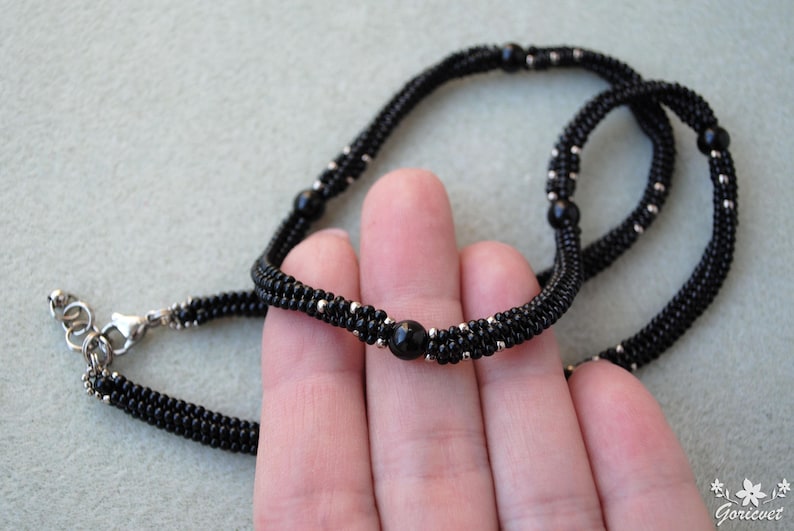 May include: A black beaded necklace with silver accents and a central black bead. The necklace is made of small, tightly woven beads, creating a textured appearance. The clasp is silver. The jewelry is displayed against a light gray background.