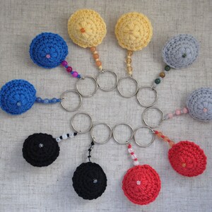 May include: A set of ten crocheted keyrings in various colours, including red, blue, yellow, black, and grey. Each keyring features a small crocheted ball with a metal ring attached. The keyrings are arranged in a circle on a textured surface.