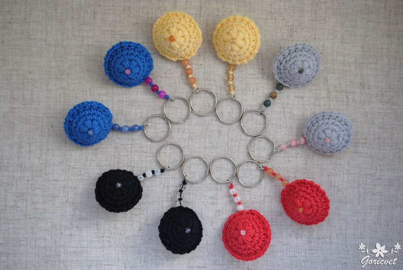 May include: A set of ten crocheted keyrings in various colours, including red, blue, yellow, black, and grey. Each keyring features a small crocheted ball with a metal ring attached. The keyrings are arranged in a circle on a textured surface.