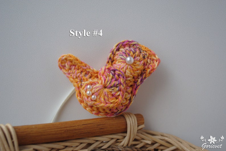 May include: A crocheted bird with a swirl pattern in shades of orange, pink, and purple. The bird has three white pearl beads for eyes. The bird is sitting on a wicker basket.