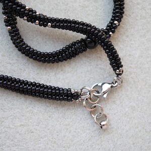 May include: Close-up of a black beaded necklace with silver-colored accents and a silver clasp. The necklace is made of small, round beads and has a braided appearance. The background is a light gray.