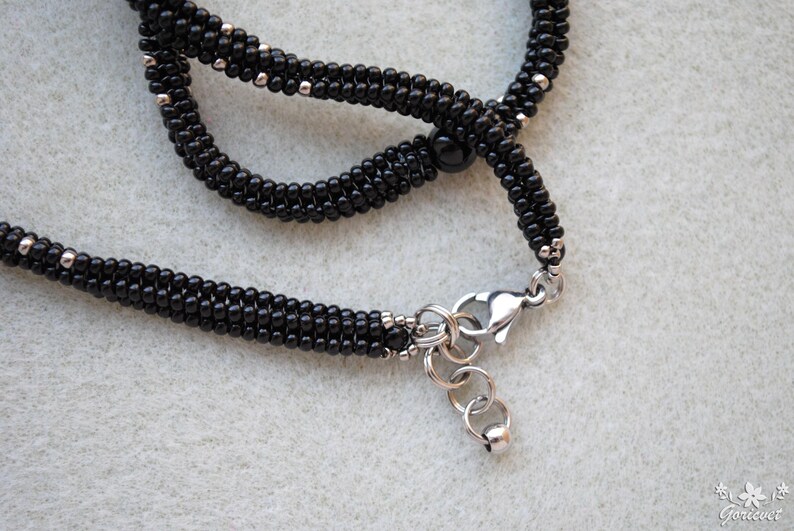 May include: Close-up of a black beaded necklace with silver-colored accents and a silver clasp. The necklace is made of small, round beads and has a braided appearance. The background is a light gray.