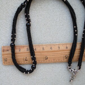 May include: A black beaded necklace with silver accents and a silver clasp. The necklace is laid out on a light gray surface next to a wooden ruler. The necklace measures approximately 13 inches in length.