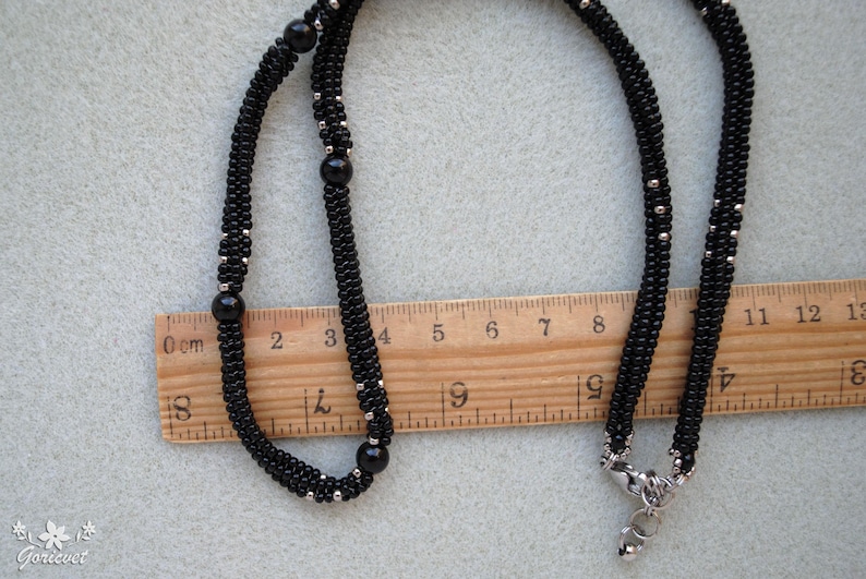 May include: A black beaded necklace with silver accents and a silver clasp. The necklace is laid out on a light gray surface next to a wooden ruler. The necklace measures approximately 13 inches in length.