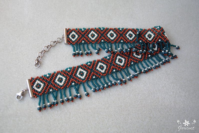 K&ouml;nnte beinhalten: Zwei Armb&auml;nder mit Perlenbesatz und geometrischem Muster. Die Armb&auml;nder weisen ein sich wiederholendes Design aus wei&szlig;en Rauten, orangefarbenen und t&uuml;rkisfarbenen Perlen auf. Die Armb&auml;nder haben einen Fransenbesatz aus t&uuml;rkisfarbenen Perlen mit kleinen schwarzen Perlen an den Enden. Die Armb&auml;nder sind mit einer silbernen Kette und einem Verschluss verbunden.