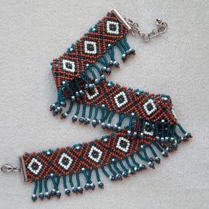K&ouml;nnte beinhalten: Ein Perlenarmband mit geometrischem Muster. Das Armband hat ein sich wiederholendes Design aus wei&szlig;en Rauten vor einem Hintergrund aus orangefarbenen und dunkelblauen Perlen. T&uuml;rkisfarbene Fransen h&auml;ngen von unten.