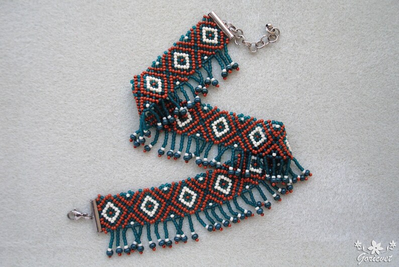 K&ouml;nnte beinhalten: Ein Perlenarmband mit geometrischem Muster. Das Armband hat ein sich wiederholendes Design aus wei&szlig;en Rauten vor einem Hintergrund aus orangefarbenen und dunkelblauen Perlen. T&uuml;rkisfarbene Fransen h&auml;ngen von unten.
