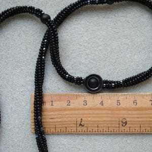 May include: A black beaded necklace with a black metal pendant. The necklace is measured against a wooden ruler with markings in inches.