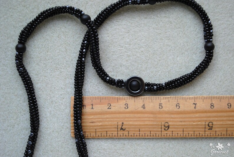 May include: A black beaded necklace with a black metal pendant. The necklace is measured against a wooden ruler with markings in inches.