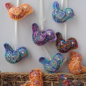 May include: A collection of colorful crocheted bird ornaments with pearl accents. The birds are hanging from white string and are arranged in a basket.