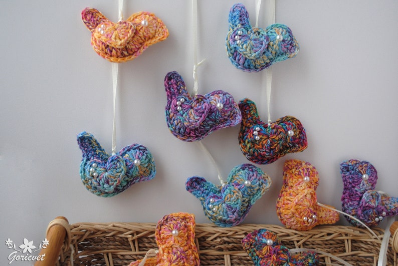 May include: A collection of colorful crocheted bird ornaments with pearl accents. The birds are hanging from white string and are arranged in a basket.