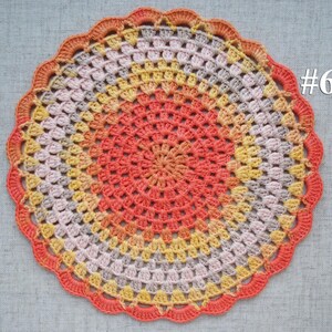 Puede incluir: Un tapete redondo hecho a mano con ganchillo en tonos naranja, amarillo y gris. El tapete presenta un borde festoneado y un dise&ntilde;o central que irradia hacia afuera. El n&uacute;mero "#6" es visible en la esquina superior derecha.