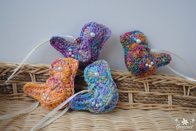 May include: Four crocheted bird ornaments in different colors with pearl beads. The ornaments are displayed on a wicker basket.