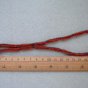 May include: A necklace made of small, reddish-orange beads, with a silver clasp. The necklace is laid out on a light gray surface next to a wooden ruler, measuring approximately 19 inches in length.