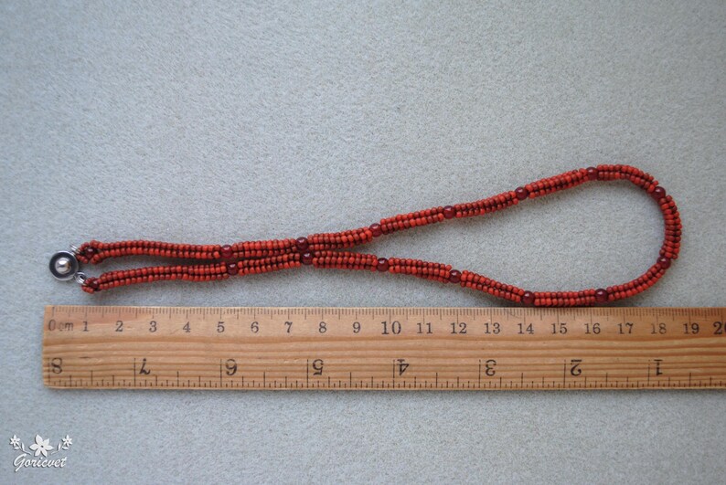 May include: A necklace made of small, reddish-orange beads, with a silver clasp. The necklace is laid out on a light gray surface next to a wooden ruler, measuring approximately 19 inches in length.