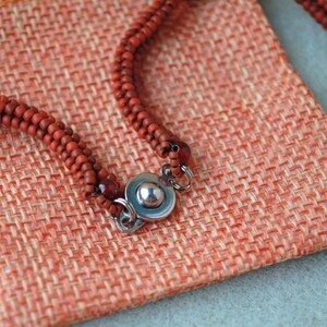 May include: Close-up of a reddish-brown beaded necklace with a silver clasp, resting on a textured orange fabric pouch. The beads are small and evenly spaced. The pouch has a woven appearance.