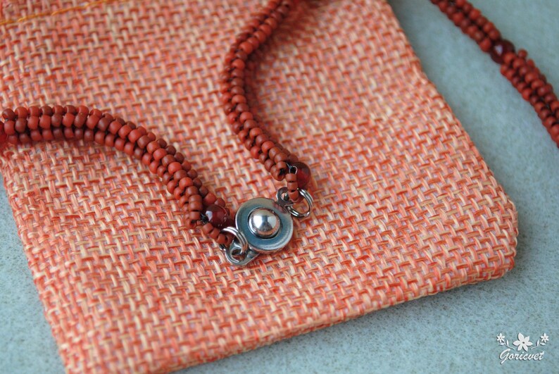 May include: Close-up of a reddish-brown beaded necklace with a silver clasp, resting on a textured orange fabric pouch. The beads are small and evenly spaced. The pouch has a woven appearance.