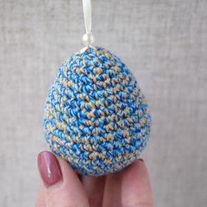 Pu&ograve; includere: Un ornamento pasquale a forma di uovo in crochet blu e beige con un nastro bianco.