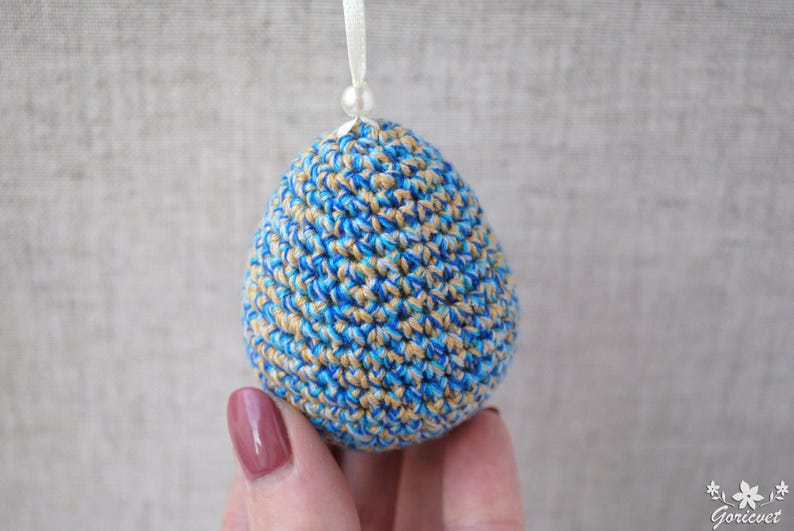 Pu&ograve; includere: Un ornamento pasquale a forma di uovo in crochet blu e beige con un nastro bianco.