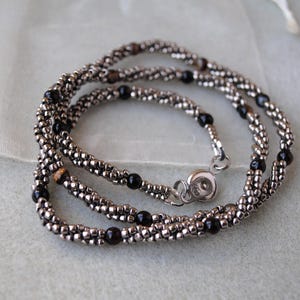 Op de afbeelding: Een zilveren en zwarte kralenketting met een magneetsluiting. De ketting is gemaakt van kleine, ronde kralen en heeft een gelaagd ontwerp.