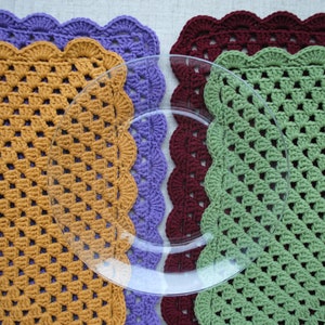May include: Three crocheted placemats in different colors: mustard yellow, purple, and green. Each placemat has a scalloped edge and a square shape. A clear glass plate is placed on top of the placemats.