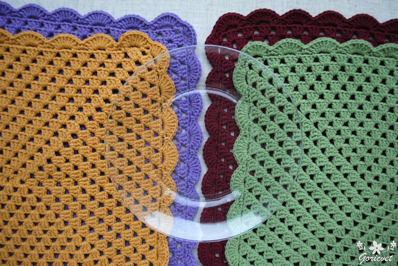 May include: Three crocheted placemats in different colors: mustard yellow, purple, and green. Each placemat has a scalloped edge and a square shape. A clear glass plate is placed on top of the placemats.