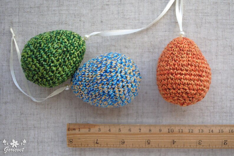Pu&ograve; includere: Tre uova di Pasqua all'uncinetto appese a un nastro bianco. Le uova sono verdi, blu e arancioni con un motivo maculato.
