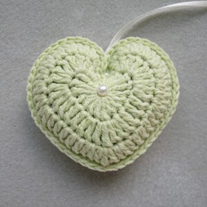 May include: A light green crocheted heart with a white pearl in the centre. The heart is attached to a white ribbon.