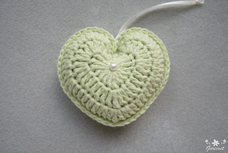 May include: A light green crocheted heart with a white pearl in the centre. The heart is attached to a white ribbon.
