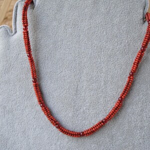 May include: A red beaded necklace displayed on a gray, felt-like surface. The necklace is composed of small, round beads and features a twisted design. The beads are a vibrant red color, with darker red accents interspersed throughout.