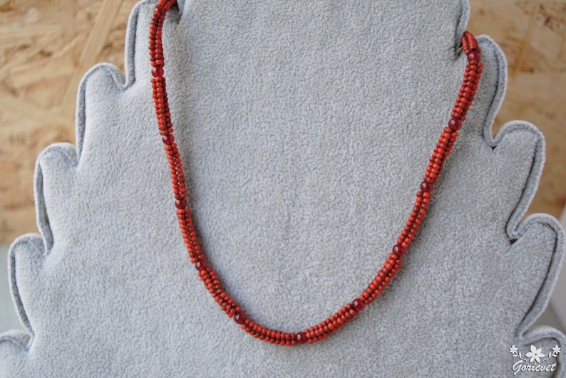 May include: A red beaded necklace displayed on a gray, felt-like surface. The necklace is composed of small, round beads and features a twisted design. The beads are a vibrant red color, with darker red accents interspersed throughout.