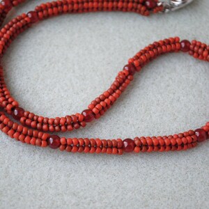 May include: A handmade necklace featuring multiple strands of small, vibrant red beads. The necklace has a braided appearance with larger, darker red beads interspersed throughout. A silver clasp secures the necklace.