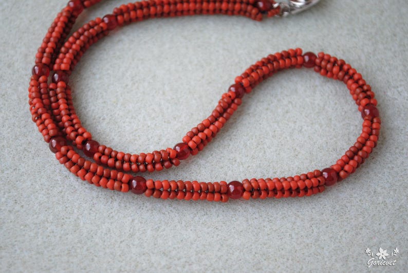 May include: A handmade necklace featuring multiple strands of small, vibrant red beads. The necklace has a braided appearance with larger, darker red beads interspersed throughout. A silver clasp secures the necklace.
