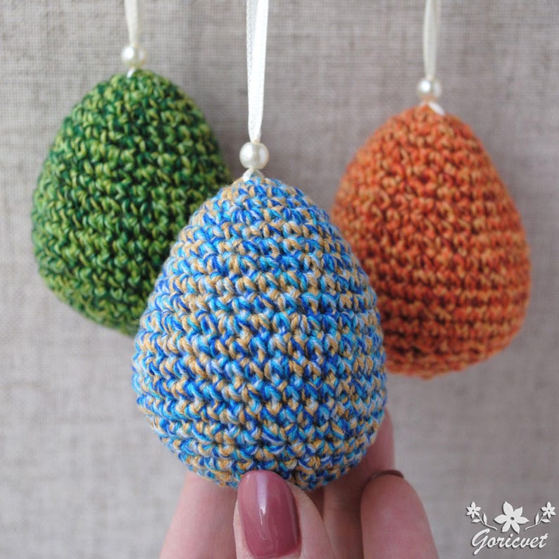 Pu&ograve; includere: Tre uova di Pasqua all'uncinetto in verde, blu e arancione. Le uova sono appese a uno spago bianco.