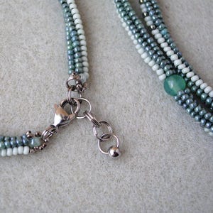 May include: A multi-strand necklace with a silver clasp and a green gemstone bead. The necklace is made of small, light blue, white, and dark blue beads.