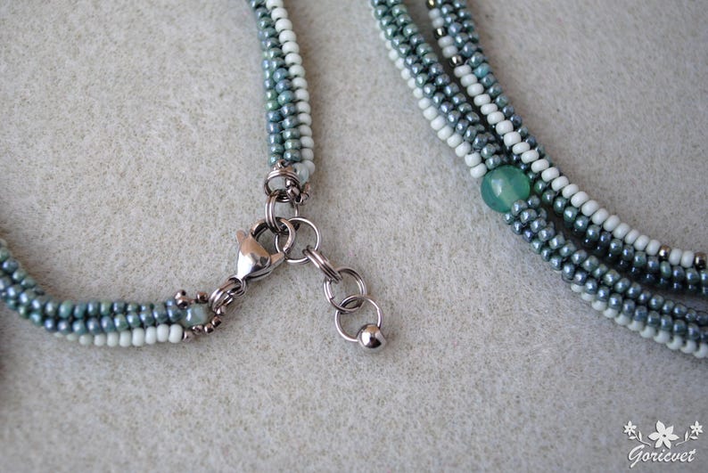 May include: A multi-strand necklace with a silver clasp and a green gemstone bead. The necklace is made of small, light blue, white, and dark blue beads.