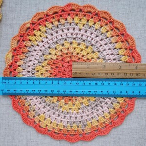 Puede incluir: Un tapete de ganchillo hecho a mano y colorido con un borde festoneado. El tapete presenta un degradado de colores, incluyendo naranja, amarillo y rosa. El di&aacute;metro es de aproximadamente 25 cm. Una regla se coloca sobre el tapete para medir.