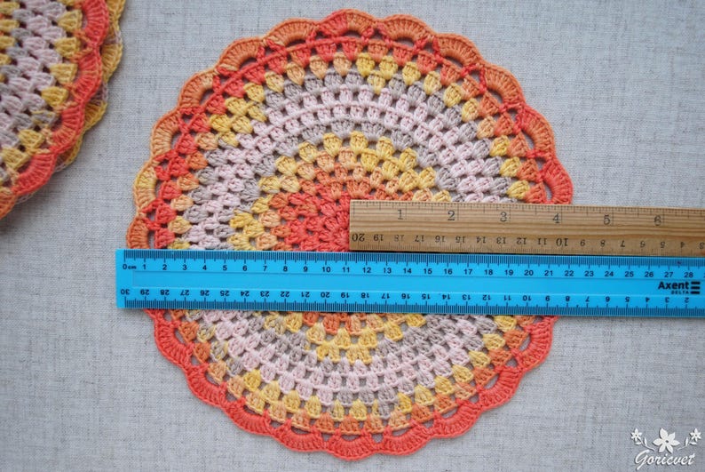 Puede incluir: Un tapete de ganchillo hecho a mano y colorido con un borde festoneado. El tapete presenta un degradado de colores, incluyendo naranja, amarillo y rosa. El di&aacute;metro es de aproximadamente 25 cm. Una regla se coloca sobre el tapete para medir.