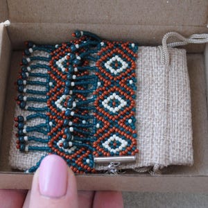 K&ouml;nnte beinhalten: Handgefertigtes Perlenarmband in einer Pappschachtel. Das Armband hat ein geometrisches Muster mit teal, orange und wei&szlig;en Perlen. Es wird mit einem kleinen beigen Kordelzugbeutel geliefert. Das Armband wird mit einem silbernen Verschluss gesichert.