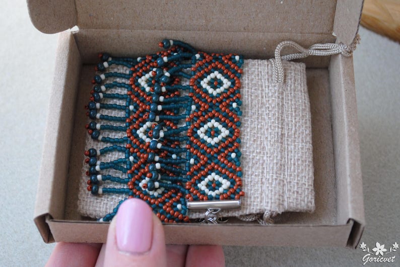 K&ouml;nnte beinhalten: Handgefertigtes Perlenarmband in einer Pappschachtel. Das Armband hat ein geometrisches Muster mit teal, orange und wei&szlig;en Perlen. Es wird mit einem kleinen beigen Kordelzugbeutel geliefert. Das Armband wird mit einem silbernen Verschluss gesichert.