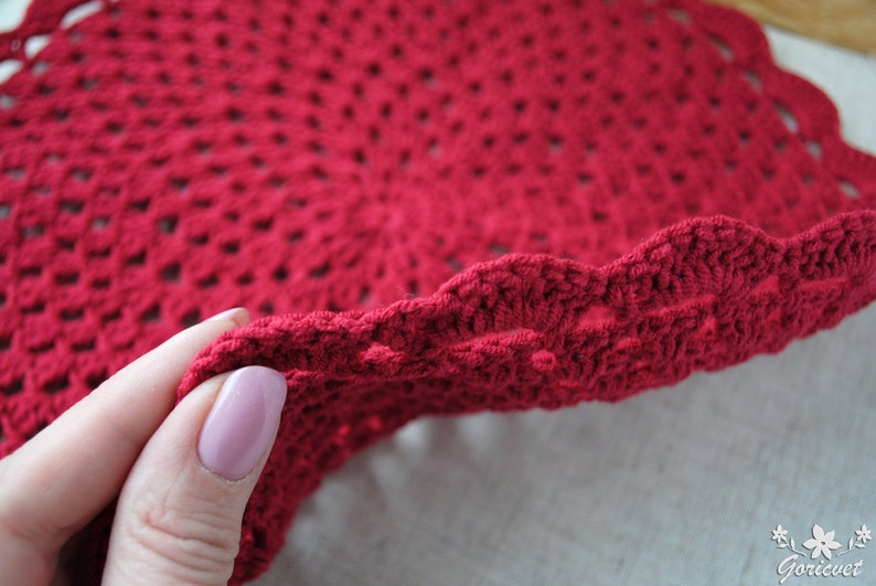 Pu&ograve; includere: Primo piano di un centrino all'uncinetto rosso con bordo a festoni. Il centrino &egrave; tenuto da una mano.
