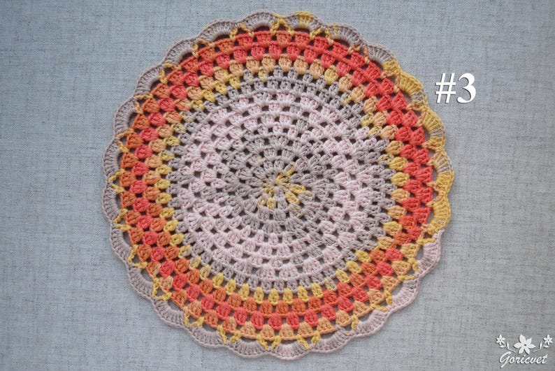 Puede incluir: Un tapete de ganchillo circular hecho a mano en tonos melocot&oacute;n, amarillo y gris. El tapete presenta un borde festoneado y un dise&ntilde;o central en espiral. El n&uacute;mero "#3" es visible en la esquina superior derecha.