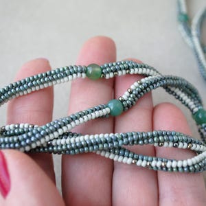 May include: A multi-strand necklace made with small, gray, white, and green beads. The necklace is made with a variety of beads, including round, oval, and square beads. The necklace is strung on a silver chain.